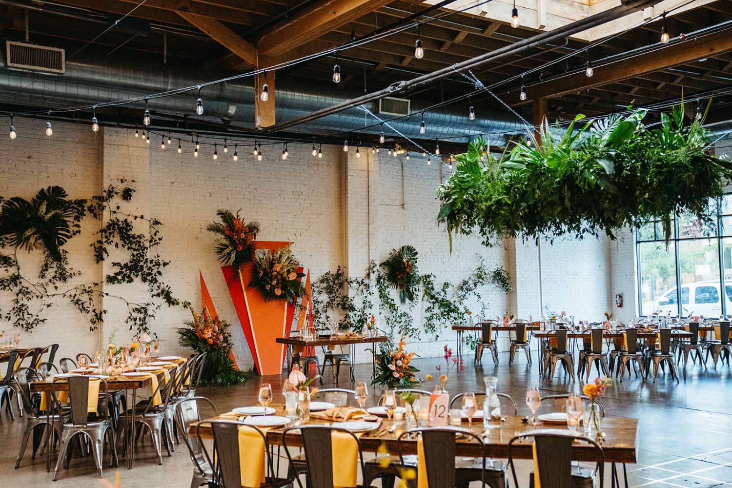 Wedding reception set-up with tables and chairs, greenery chandeliers, and a geometric orange backdrop behind the sweetheart table