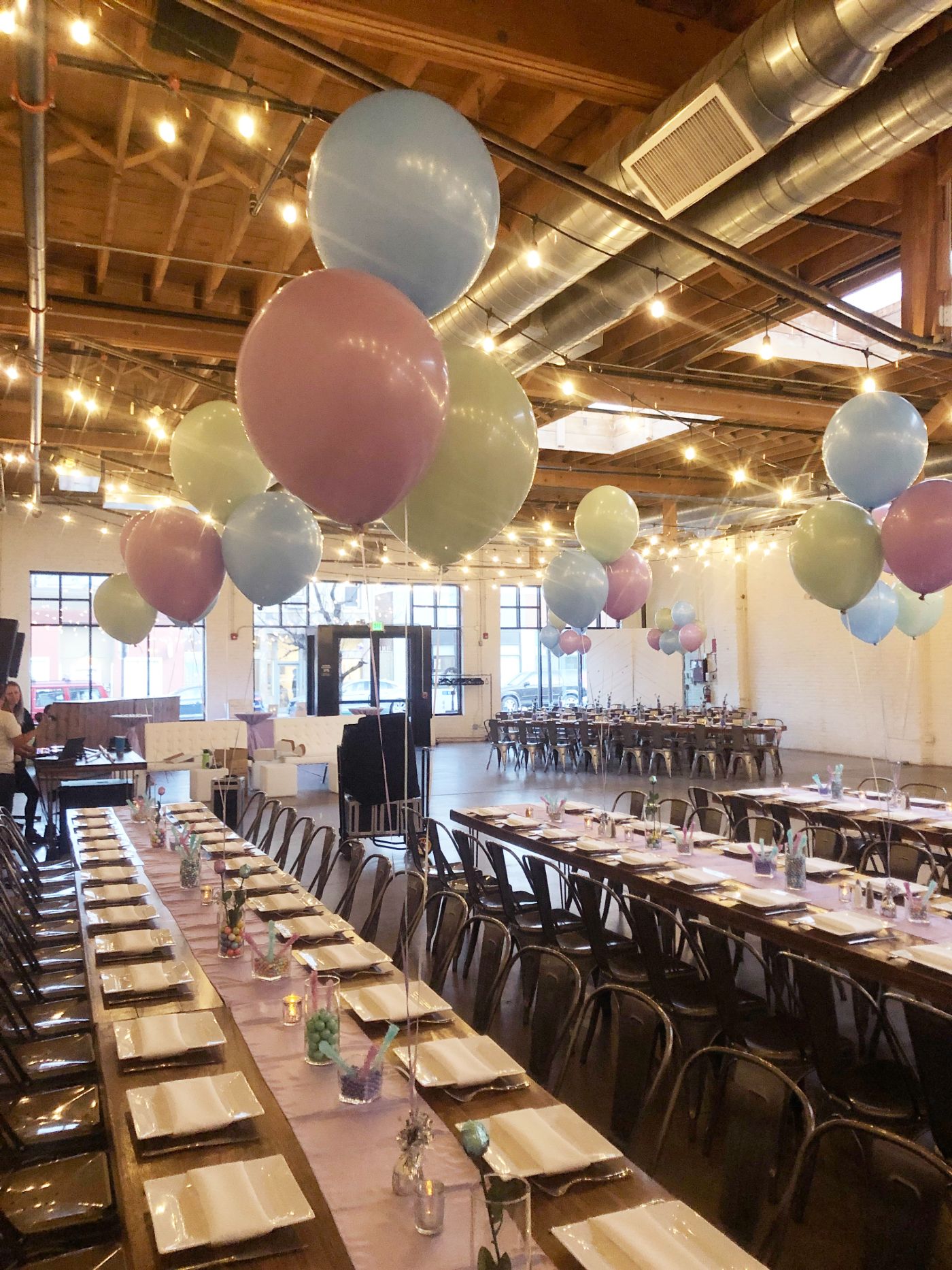 Pastel-themed event setup inside an industrial-style venue with high ceilings, exposed beams, and warm string lights. Long wooden banquet tables are decorated with lavender runners, white plates, and colorful candy centerpieces, complemented by oversized pastel balloons in blue, pink, and mint green. Perfect for baby showers, kids’ parties, or whimsical celebrations.