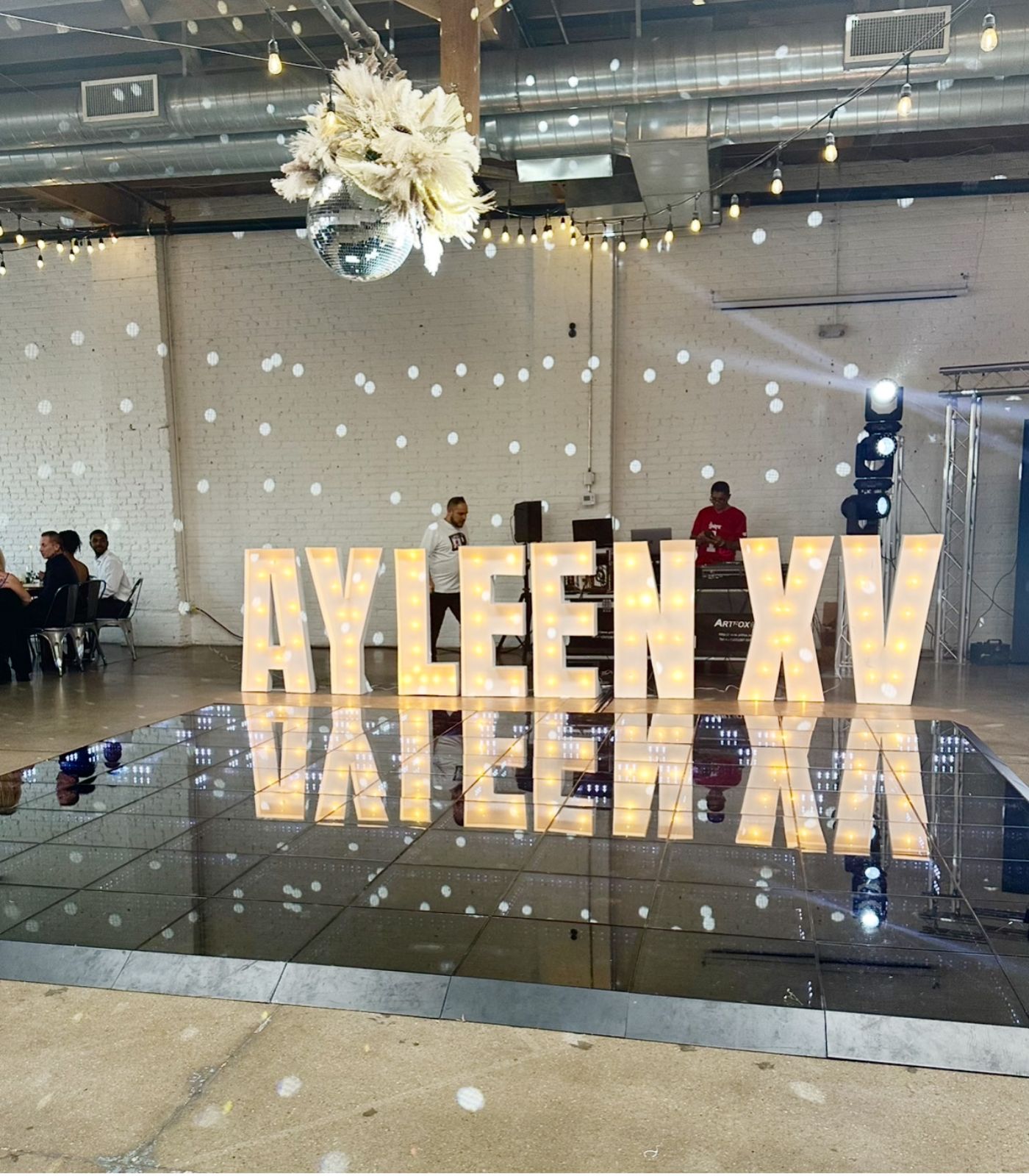 Illuminated marquee letters spelling on a shiny black dance floor, reflecting the letters. A disco ball hangs from the ceiling, casting light patterns. In the background, two DJs stand behind equipment, and some guests are seated. The venue has an industrial-chic feel with white brick walls and exposed ceilings, suggesting an event space or boutique.