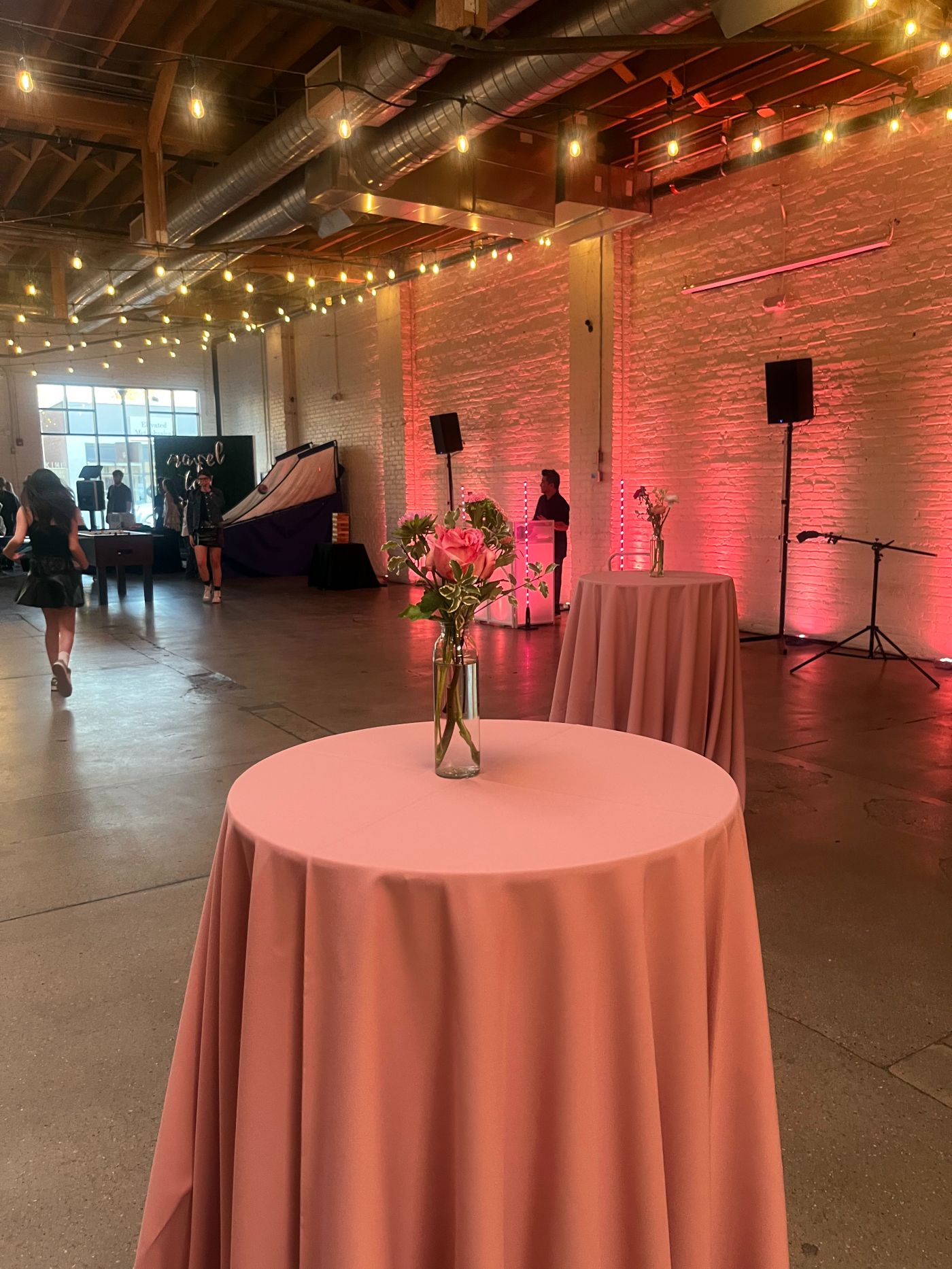 Modern event space featuring pink cocktail tables with simple floral centerpieces and soft pink uplighting against white brick walls. Exposed ductwork and string lights add an urban-industrial charm, ideal for wedding receptions, networking events, or social mixers.