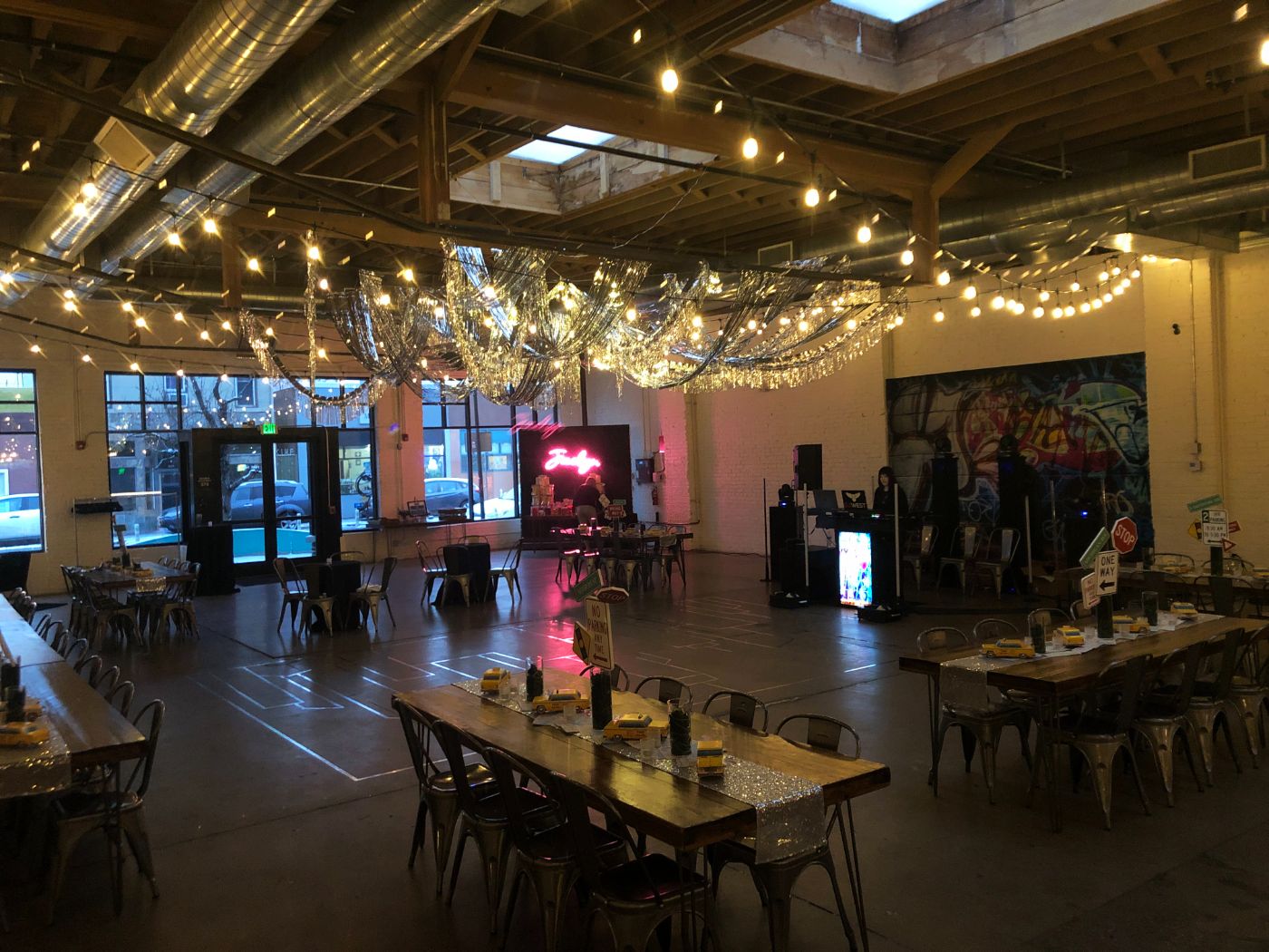 Trendy event venue with dramatic silver tinsel ceiling decor, neon signage, and graffiti-style mural. Wooden tables with casual yellow-themed place settings and a DJ booth create a fun, modern atmosphere for birthdays, themed parties, or teen celebrations in a warehouse-style space.