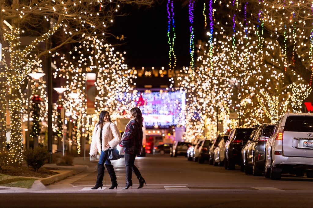 Two women crossing the street in the middle of a holiday market surrounded by holiday lights