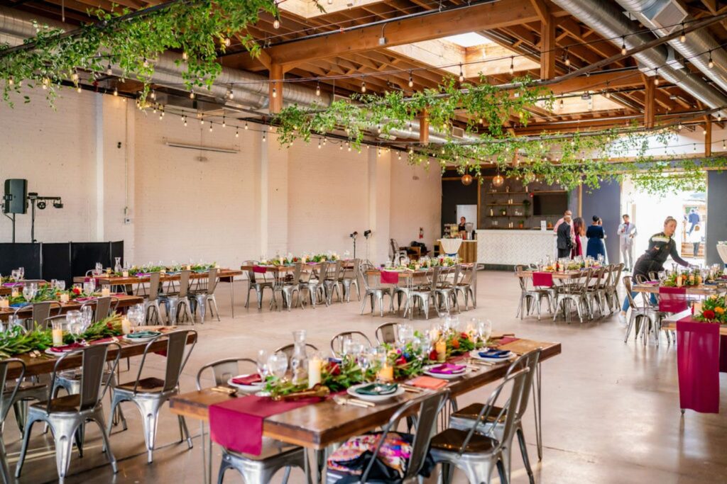 Wedding reception at SKYLIGHT in Denver featuring wooden farm tables, colorful place settings, and greenery strung from the ceiling.