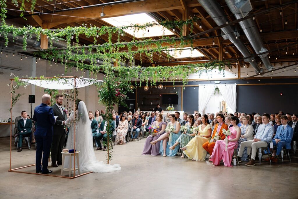 Colorful wedding ceremony at SKYLIGHT in Denver, CO, with a lush greenery ceiling installation and vibrant bridal party attire.