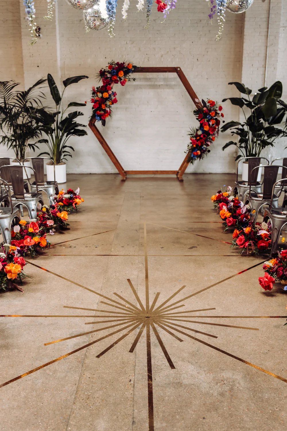 Colorful wedding ceremony at SKYLIGHT Denver with vibrant pink and orange florals lining the aisle, a wooden backdrop with matching florals, and a gold mylar tape geometric design